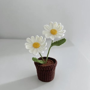 Op de afbeelding: Een gehaakte potplant met twee witte madeliefjes en groene bladeren. De pot is bruin en heeft een getextureerd oppervlak.