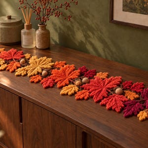 May include: A long, crocheted table runner featuring a dense arrangement of autumn-colored leaves in shades of deep red, bright orange, and golden yellow, interspersed with small, round, light brown crocheted acorns throughout the design.