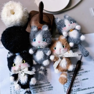 May include: Five crocheted cat toys, each with a different color and pattern. The cats are all sitting on a white surface with a piece of paper and a wooden yarn holder.