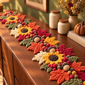May include: A long, decorative table runner featuring a crochet design of autumn-themed elements including yellow sunflowers, red and orange maple leaves, green foliage, small berries, and tan acorns arranged in a repeating pattern.