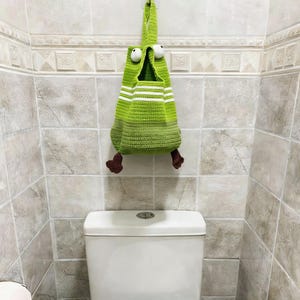 Può includere: Un sacco portaoggetti a forma di rana all'uncinetto verde fatto a mano con strisce bianche e grandi occhi bianchi. La borsa ha zampe marroni ed è appesa a un gancio sopra un water bianco. Il muro del bagno è piastrellato.