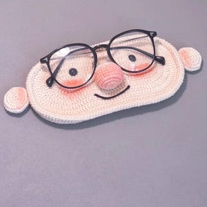Könnte beinhalten: Eine gehäkelte Brillenhülle in Form eines lächelnden Gesichts mit rosa Nase und Wangen. Die Hülle enthält eine Brille mit schwarzem Gestell.