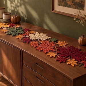 May include: A long, decorative table runner composed of interconnected, crocheted maple leaves in a vibrant autumn color palette of deep burgundy, burnt orange, golden yellow, and forest green, arranged in a dense, overlapping pattern.