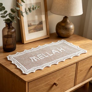 Puede incluir: Un tapete rectangular de ganchillo blanco con un borde festoneado y la palabra MELLAH tejida en el centro. La pieza está elaborada con un patrón de rejilla fina y se muestra extendida, destacando el intrincado trabajo y la textura.
