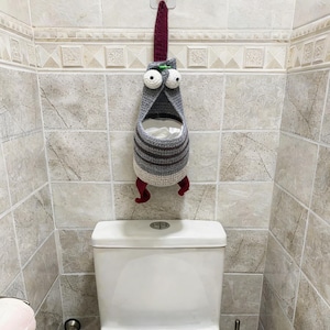 Può includere: Un portarotolo di carta igienica all'uncinetto fatto a mano a forma di creatura. Il supporto è grigio con strisce marroni, grandi occhi mobili e piedi rossi. È appeso a un gancio sopra un water bianco, aggiungendo un tocco giocoso all'arredamento del bagno.