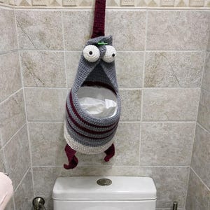Può includere: Un portarotolo di carta igienica all'uncinetto fatto a mano e stravagante a forma di personaggio dei cartoni animati. Il supporto è grigio con strisce bordeaux e dettagli bianchi, con grandi occhi bianchi e piedi bordeaux. È appeso a un gancio a parete.