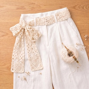 May include: Off-white linen trousers featuring a wide, intricate crochet lace belt tied in a bow at the waist. A ball of matching off-white crochet yarn with a wooden crochet hook inserted through the center sits on the fabric of the trousers.