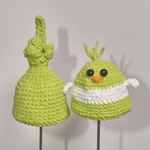 May include: Two handmade crochet hats in bright green. One hat is shaped like a finger with a knot at the top. The other hat is shaped like a bird with a white stripe, black eyes, and an orange beak. Both hats are on metal rods.
