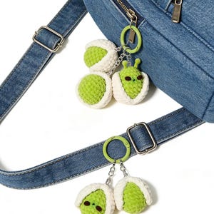 May include: Close-up of a denim backpack with a silver zipper and a green and white crochet alien keychain. The keychain features a green alien with white accents and a green ring. The backpack has a denim strap with a silver buckle.