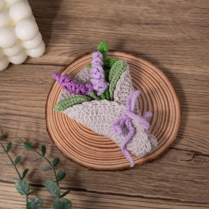 May include: A handmade crochet bouquet featuring lavender and green leaves, wrapped in a light gray crocheted cone and tied with a lavender ribbon. The bouquet is displayed on a wooden slice, showcasing the intricate details of the craft.