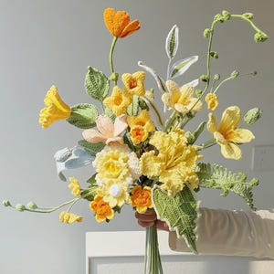 May include: A crocheted bouquet of yellow and orange flowers with green leaves. The flowers are arranged in a hand-tied bouquet.