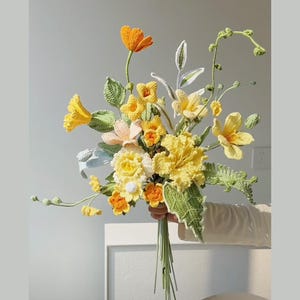 May include: A crocheted bouquet of yellow and orange flowers with green leaves. The bouquet is held in a hand.