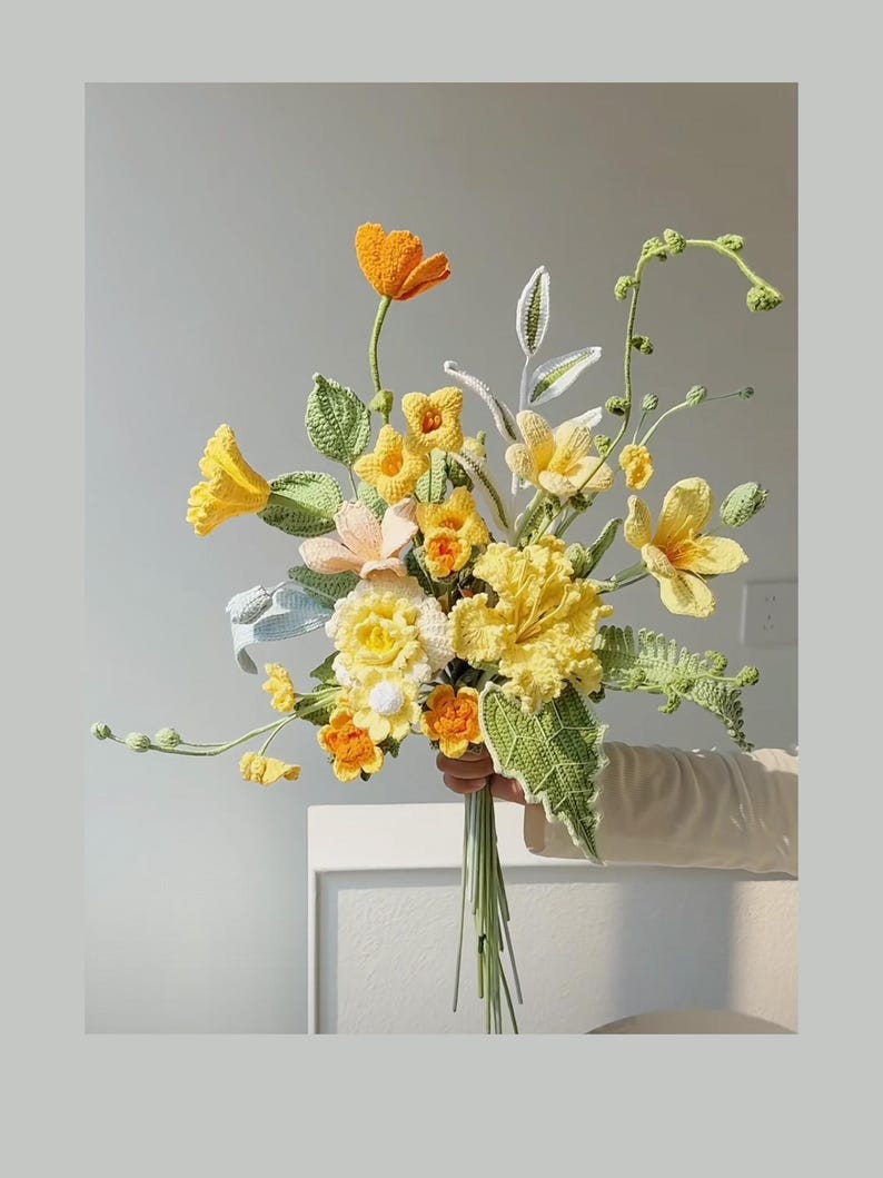 May include: A crocheted bouquet of yellow and orange flowers with green leaves. The bouquet is held in a hand.