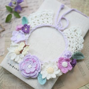 Puede incluir: Una guirnalda de flores de ganchillo con un dije de mariposa. La guirnalda está hecha de hilo blanco, morado, rosa y azul claro. Está atada con una cinta morada.