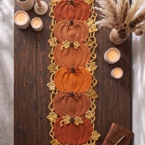 Peut inclure: Un chemin de table rectangulaire composé de six citrouilles crochetées dans des tons orange variés, disposées verticalement. Les citrouilles sont reliées par une bordure de feuilles et de vignes crochetées en jaune, formant un textile décoratif.