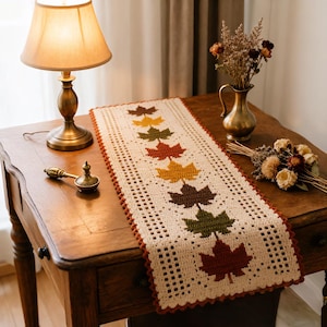 May include: A rectangular crochet table runner in cream yarn featuring a central vertical row of seven maple leaf motifs in shades of brown, yellow, orange, and green, with a scalloped border and an openwork grid pattern along the sides.