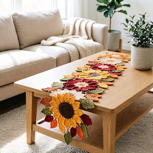 Könnte beinhalten: Ein langer, dekorativer Tischläufer mit einer Reihe gehäkelter Sonnenblumen und herbstfarbener Blätter in den Tönen Dunkelrot, Orange, Gelb und Grün. Das Blumenarrangement ist aus strukturiertem Garn gefertigt und liegt flach auf einem Holztisch.