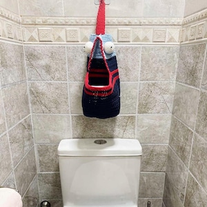 Può includere: Un portarotolo di carta igienica all'uncinetto fatto a mano e stravagante. Il supporto è blu navy con strisce e finiture rosse e ha grandi occhi mobili. È appeso a una cinghia rossa attaccata a un gancio a muro, sopra un water bianco.