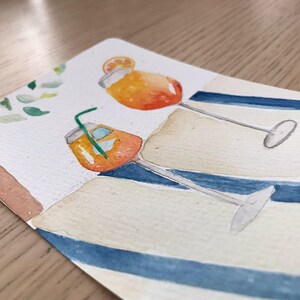 Peut inclure: Aquarelle de deux cocktails orange avec des pailles et des tranches d'orange, posés sur une surface rayée bleue et blanche. La peinture a une sensation légère et aérée, avec un arbre et des feuilles en arrière-plan.