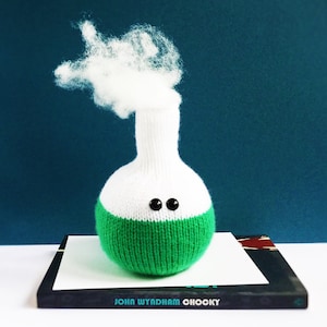 May include: A white and green knitted flask with black eyes and a white cotton cloud on top. The flask is sitting on a book with the text "John Wyndham Chocky" on the cover.