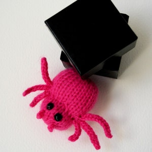 May include: A pink knitted spider toy with black eyes. The spider is sitting on top of a black box.