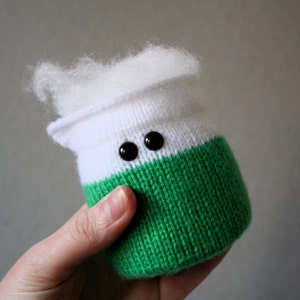 May include: A white and green knitted toy with black button eyes. The toy is shaped like a jar or container.