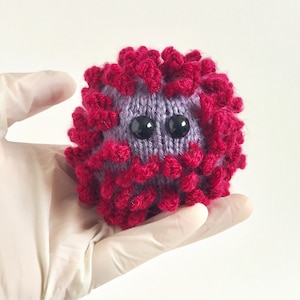 May include: A purple and red knitted plush toy shaped like a virus. The toy has two black eyes and a spiky red exterior.