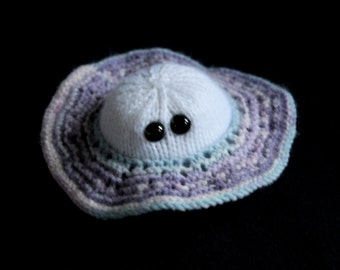 May include: A white knitted hat with two black buttons for eyes. The hat is surrounded by a purple, blue and white knitted brim.