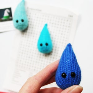 May include: Three knitted teardrop-shaped figures with black button eyes. One is held in a hand, the others are on a white surface. The figures are in shades of blue and turquoise. The figures are handmade.