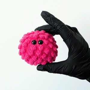 May include: A handmade, round, hot pink knitted creature with a textured surface and two black button eyes. The plush toy is held by a gloved hand against a white background. The toy has a whimsical, playful design.