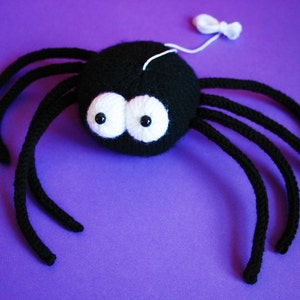 May include: A black crocheted spider with white eyes. The spider has eight legs and is sitting on a purple background.