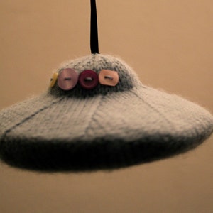 May include: A gray knitted UFO ornament with a string for hanging. The UFO has a ring of colorful buttons around the top.