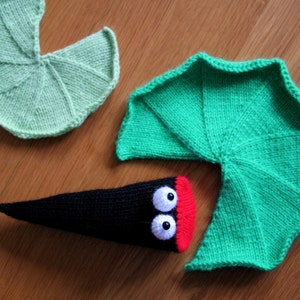 May include: A green knitted lily pad with a black knitted frog with red and white eyes. The frog is partially hidden behind the lily pad.