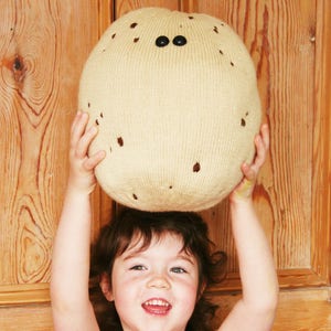 May include: A young girl holds a large, knitted, beige ball with brown spots and black eyes above her head. She is smiling and looking at the camera.