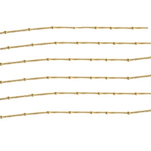 May include: Multiple strands of delicate gold-colored chain with small, evenly spaced ball beads. The chains are arranged horizontally against a white background, creating a pattern of parallel lines. The chains appear to be made of metal.