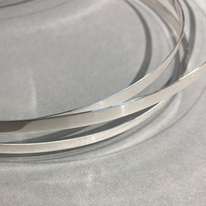 May include: Three silver metal strips, each with a slightly different width, are stacked on top of each other. The strips are curved and have a shiny, metallic finish.