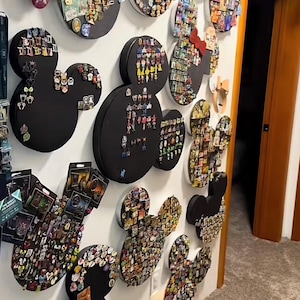 Op de afbeelding: Een verzameling Mickey Mouse-vormige prikborden aan een witte muur. Elk zwart bord is bedekt met kleurrijke spelden, waardoor een levendige weergave ontstaat. Sommige borden hebben ingelijste kunst. De kamer heeft een neutraal tapijt en een houten deur.