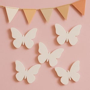 May include: Five white paper butterflies on a pink background. The butterflies are arranged in a symmetrical pattern. A string of beige and orange paper flags hangs above the butterflies.