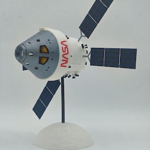 Peut inclure: Une maquette du vaisseau spatial Orion de la NASA avec un module d'équipage conique gris et un module de service blanc. Le vaisseau possède quatre ailes de panneaux solaires bleu foncé déployées et est fixé sur un support noir à base blanche.