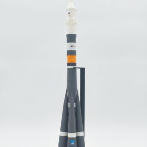 Pode incluir: Um modelo em escala de um foguete Soyuz com um esquema de cores branco, cinza e laranja. O foguete fica na vertical sobre uma base preta com a palavra SOYUZ impressa em letras maiúsculas brancas. Inclui um topo pontiagudo, seções de corpo cilíndricas e quatro propulsores cônicos na base, apoiados por um suporte vertical preto.