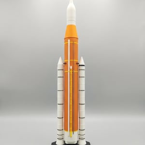 Peut inclure: Une maquette de la fusée SLS Block 1 comprenant un étage central cylindrique orange, deux propulseurs latéraux blancs et un étage supérieur blanc avec un cône de nez pointu. La fusée repose sur une base noire marquée SLS BLOCK 1.