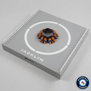 Op de afbeelding: Een vierkant grijs helipad-model met een witte cirkelvormige landingszone, witte relingen aan twee zijden en de tekst JACKLYN in het wit gedrukt. In het midden bevindt zich een klein, zeshoekig, meerkleurig mechanisch onderdeel met oranje en blauwe details.