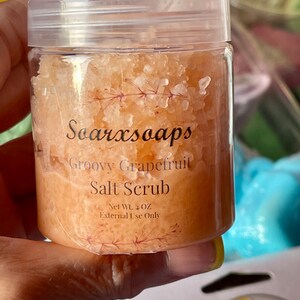 May include: A clear jar filled with a pink grapefruit salt scrub. The jar has a white lid and a label that reads "Soarxsoaps Groovy Grapefruit Salt Scrub Net Wt. 4 OZ External Use Only".