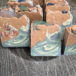 May include: Several rectangular bars of handmade soap featuring a layered design. The bottom section displays a swirling pattern of teal, blue, and white, while the top section is a solid peach color embedded with colorful flecks and smooth, translucent stones.