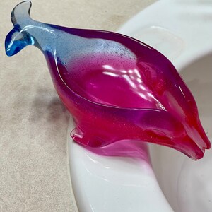 May include: A decorative, whale-shaped soap dish. The dish is made of translucent resin with a gradient of colors, transitioning from blue to purple to red. The dish is resting on a white surface.