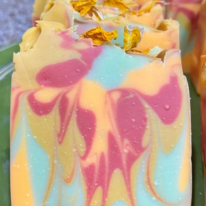 May include: A bar of handmade soap with a swirl pattern in shades of yellow, pink, and blue. The soap is decorated with dried flower petals.