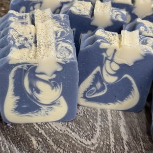 May include: Several rectangular bars of soap featuring a marbled pattern of periwinkle blue and cream white. Each bar is topped with a cream-colored star-shaped accent and a light dusting of iridescent glitter across the top surface.
