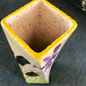 May include: A square ceramic vase with a yellow rim and a white body. The vase is decorated with purple dragonfly and black leaf designs. The interior of the vase is speckled with brown and pink spots.