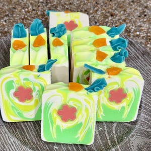 May include: Handmade soap bars with a vibrant design. The bars feature a swirl of green, yellow, and white, with a red flower, blue leaf, and orange accents. The soap is cut into rectangular shapes, perfect for a spa gift.