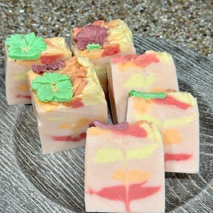 May include: Five handmade soap bars with floral designs. The soaps are layered with pink, yellow, and white swirls. Each bar has a different flower design on top, including a green flower, a pink flower, and a yellow flower.